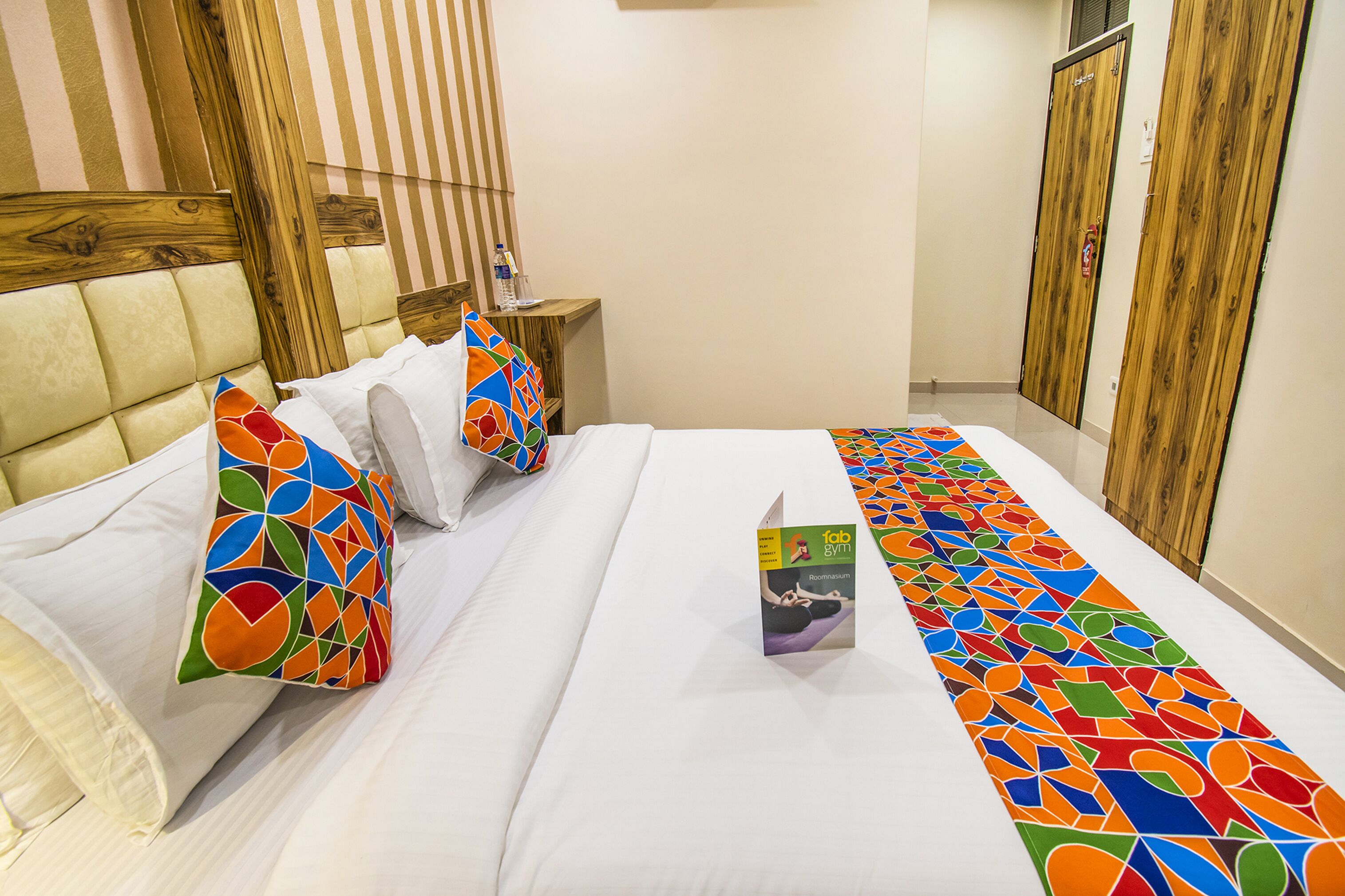 A hotel room with a large bed, a wooden headboard, a small bedside table with a water bottle, and a colorful bed runner.