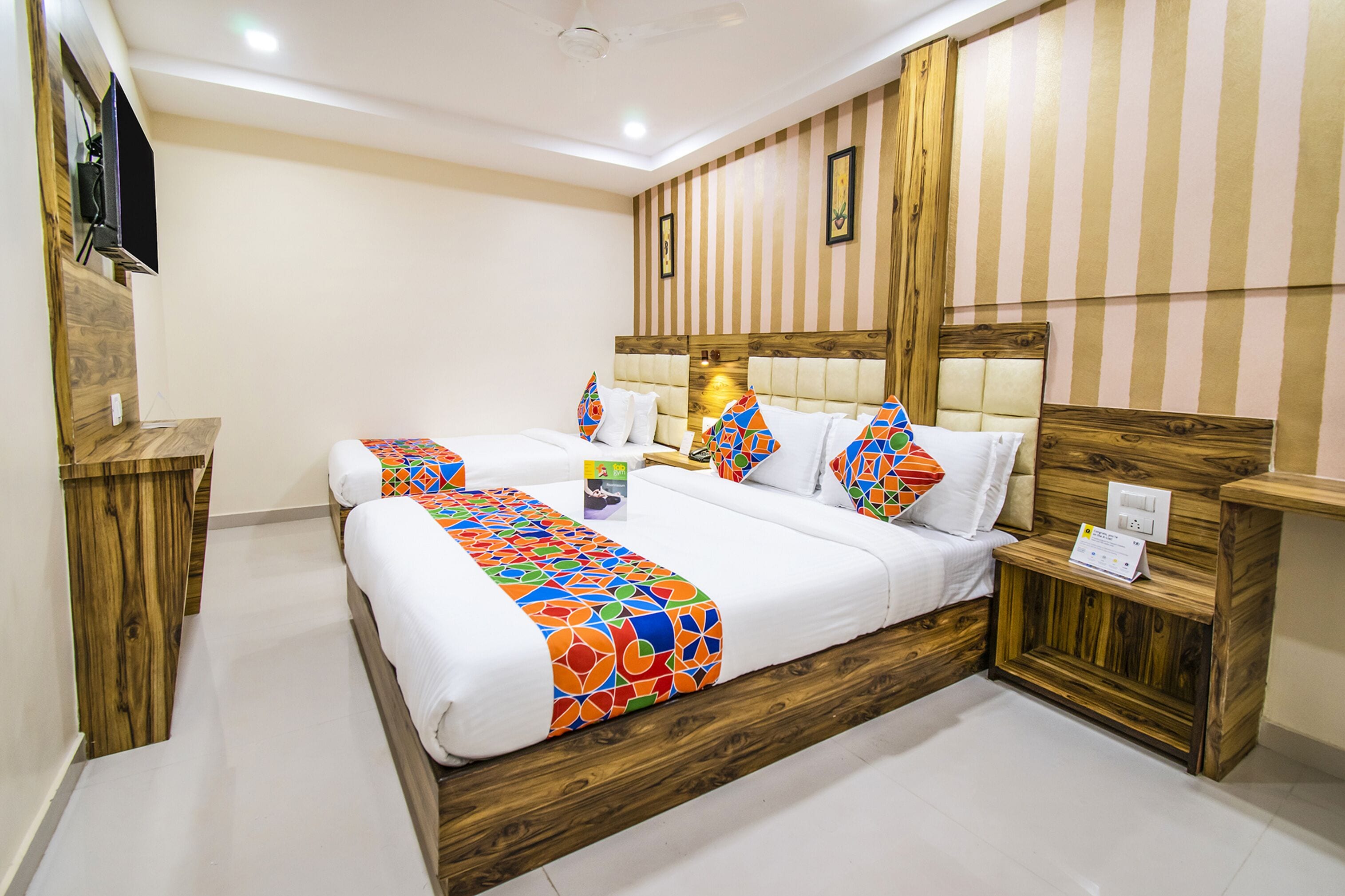 A hotel room with two beds, wooden headboards, striped wallpaper, and a flat-screen TV mounted on the wall.