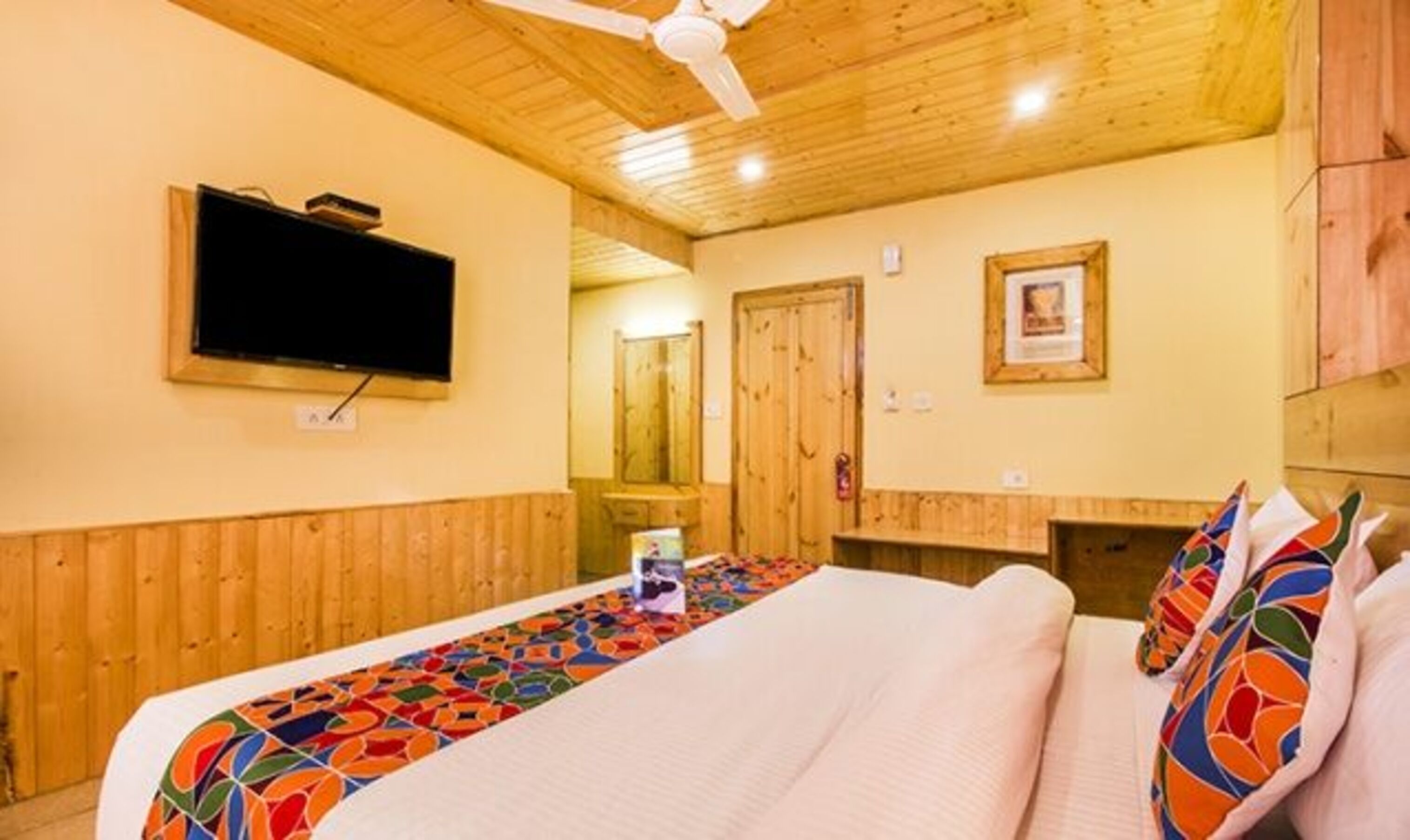 A hotel room with a wooden ceiling, a bed with a colorful bedspread, a flat-screen TV mounted on the wall, and a wooden wardrobe.