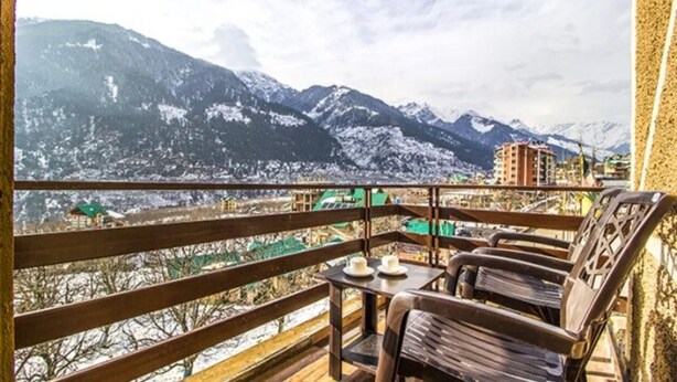 A balcony with wooden railings, a table with two chairs, and a view of snow-covered mountains and buildings. - FabHotel Shami Inn (Manali)