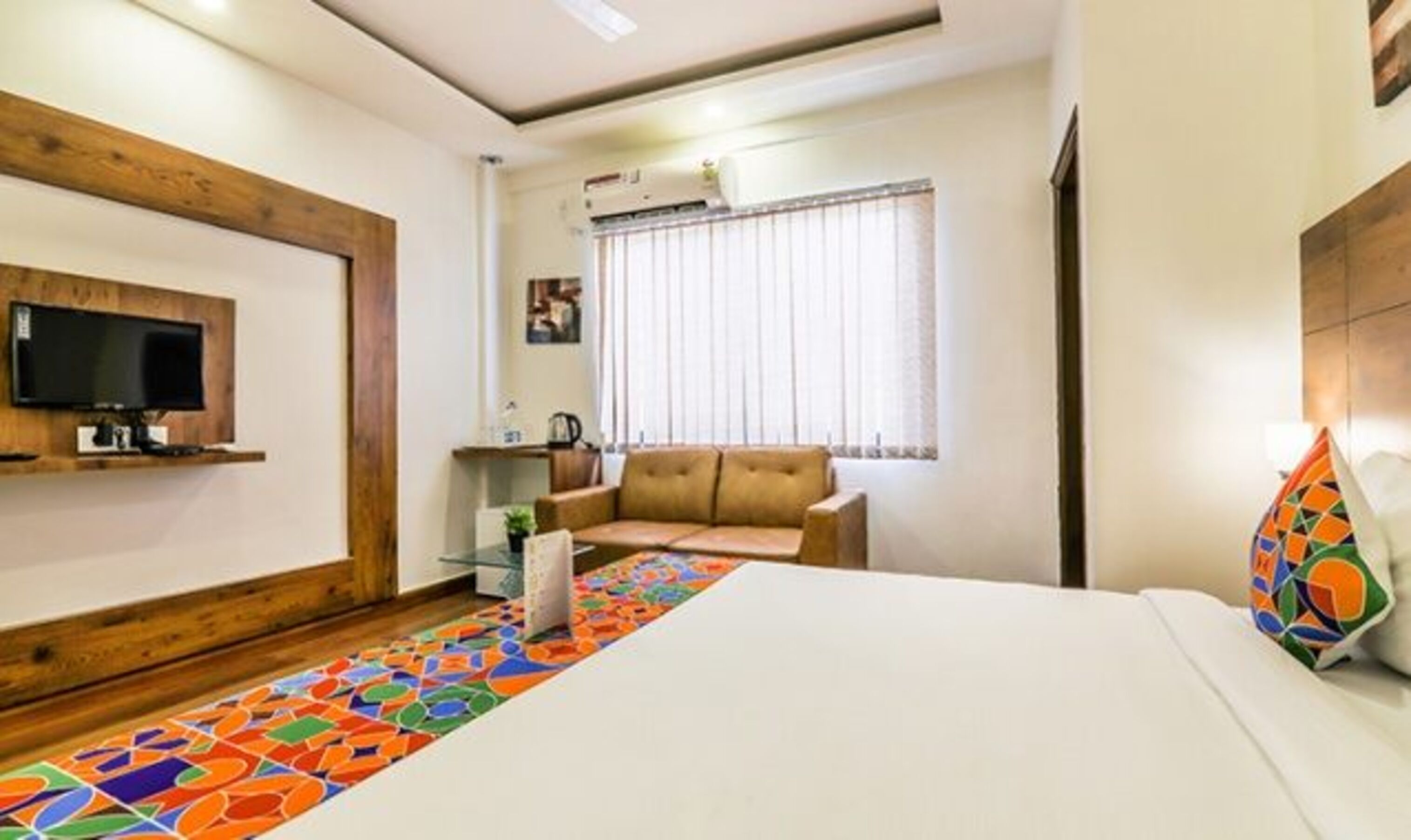A hotel room with a bed, a television mounted on the wall, a window with blinds, a sofa, and a colorful rug.