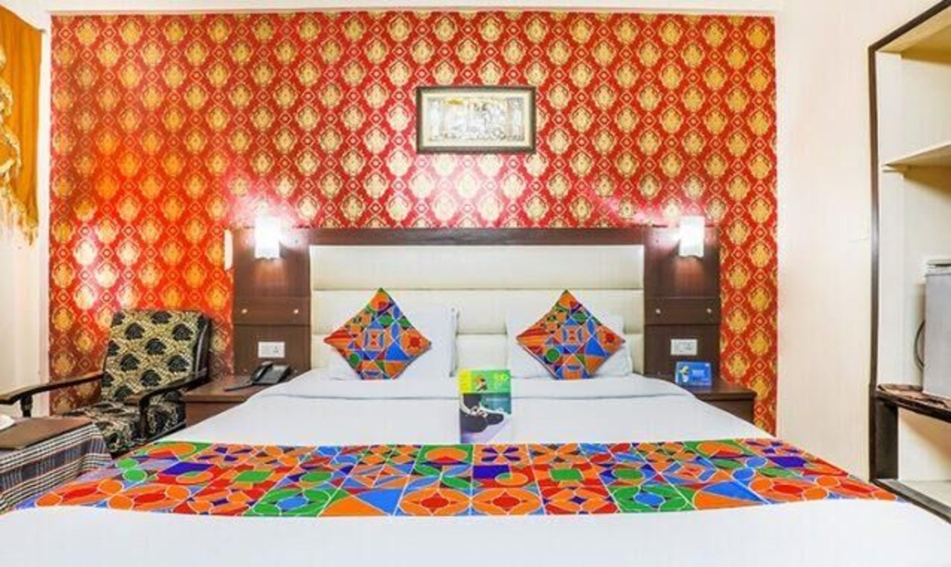 A hotel room with a patterned red and gold wallpaper, a large bed with colorful pillows, and a framed picture on the headboard.