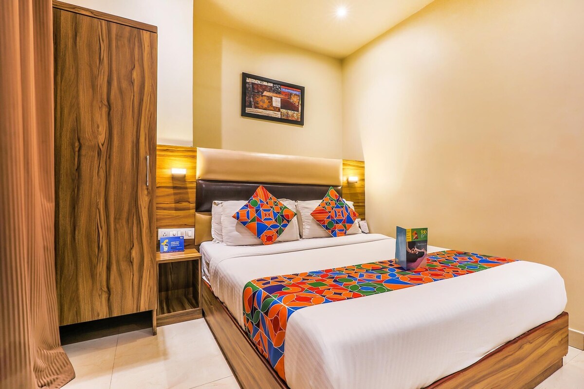 A hotel room with a wooden wardrobe, a bed with colorful bedding, a nightstand with a lamp, and a framed picture on the wall.