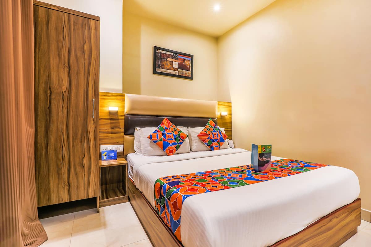 A hotel room with a wooden wardrobe, a bed with colorful bedding, a nightstand with a lamp, and a framed picture on the wall.