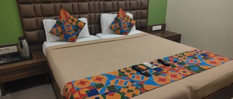 A hotel room with a brown headboard, two white pillows with colorful geometric patterns, a beige bedspread with a similar pattern, a bedside table with a kettle, and a nightstand with a remote control.
