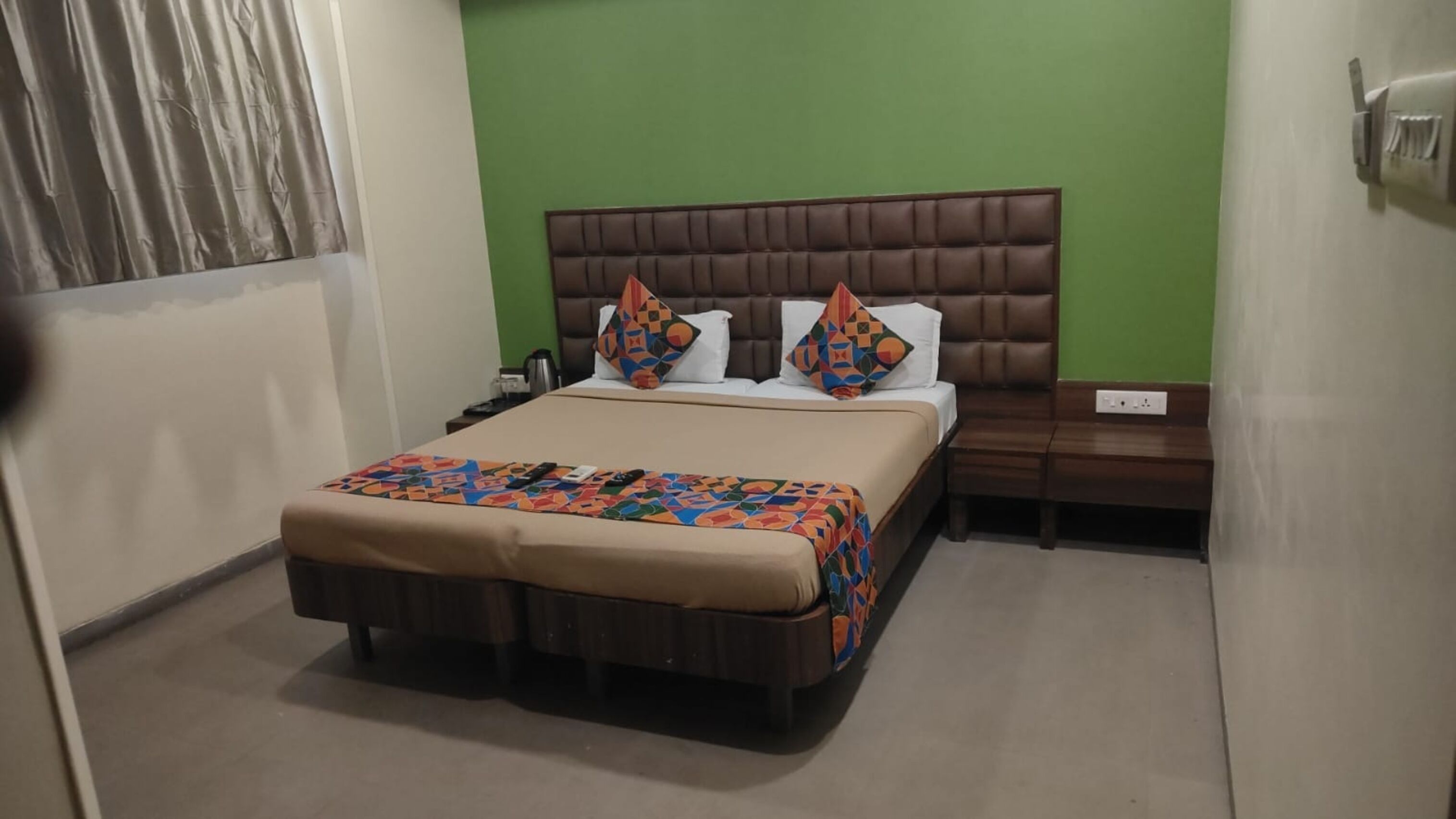 A hotel room with a large bed, a wooden nightstand, a green wall, and a window with curtains.