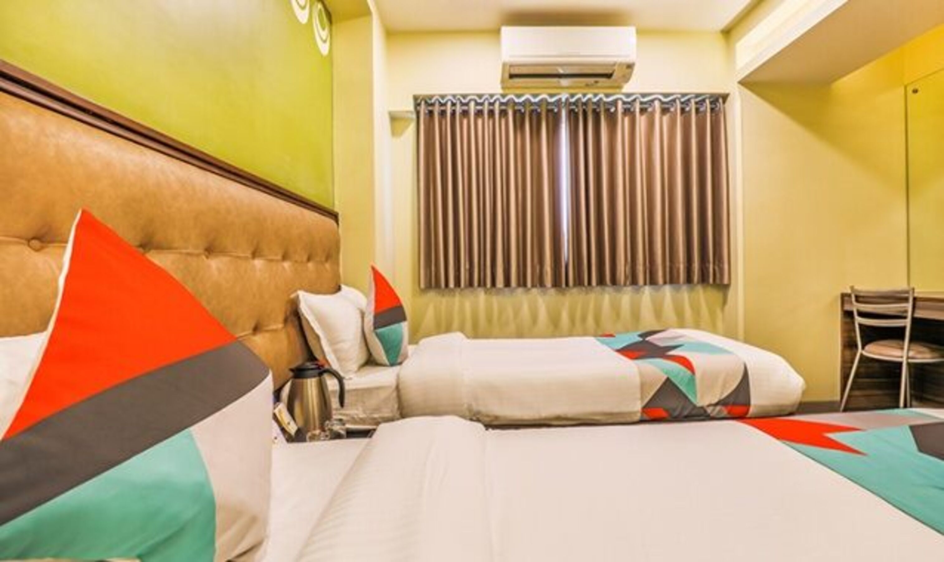 A hotel room with two beds, a headboard, a window with curtains, an air conditioning unit, and a chair.