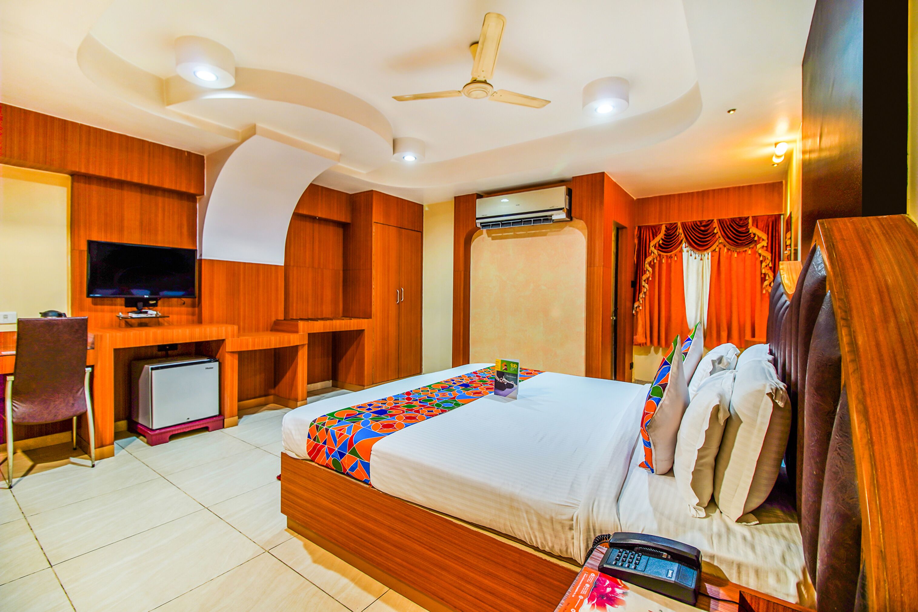 A hotel room with a bed, desk, television, air conditioning unit, and a ceiling fan.