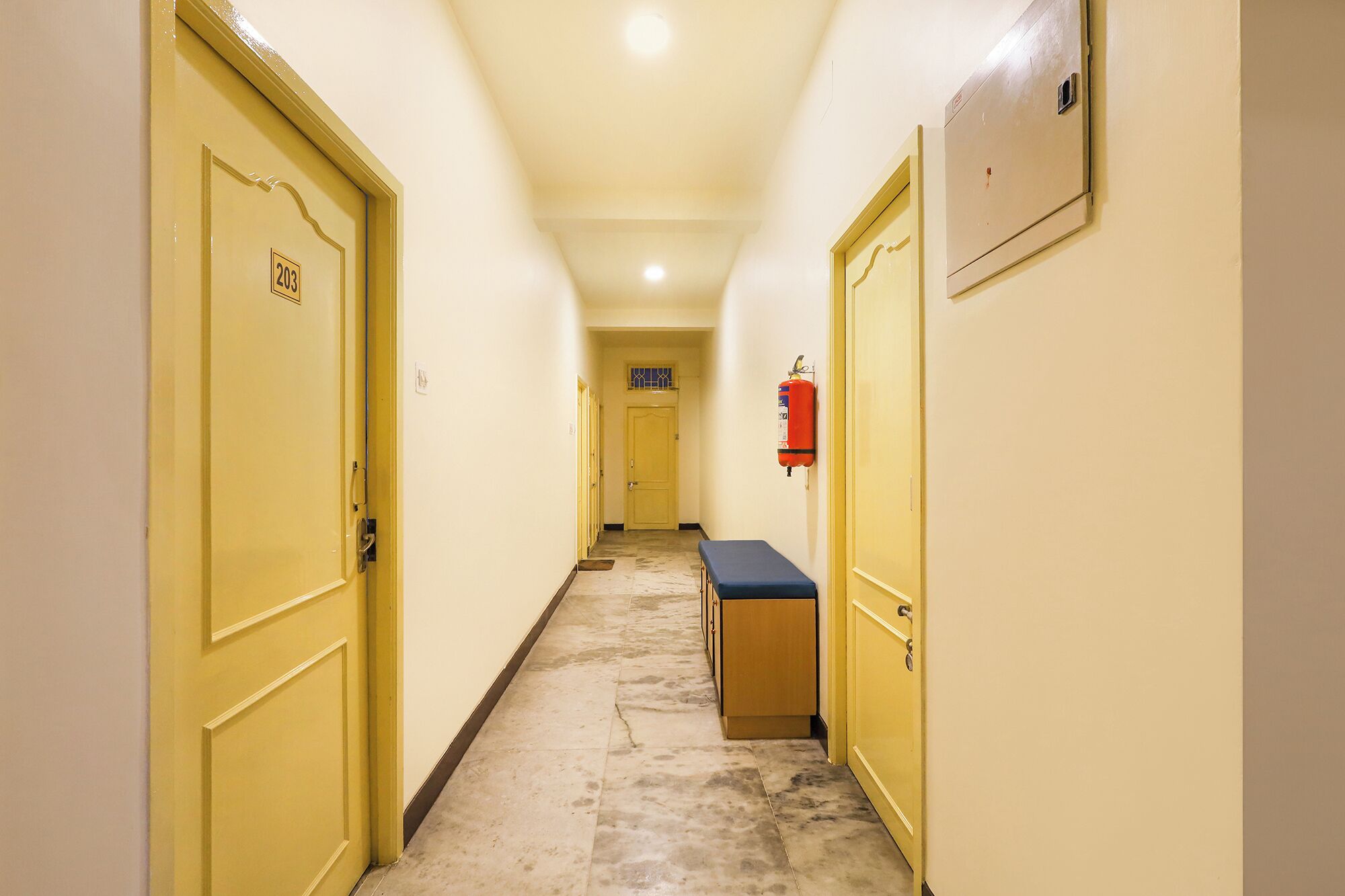 A narrow hallway with yellow doors, a fire extinguisher mounted on the wall, and a bench along one side.