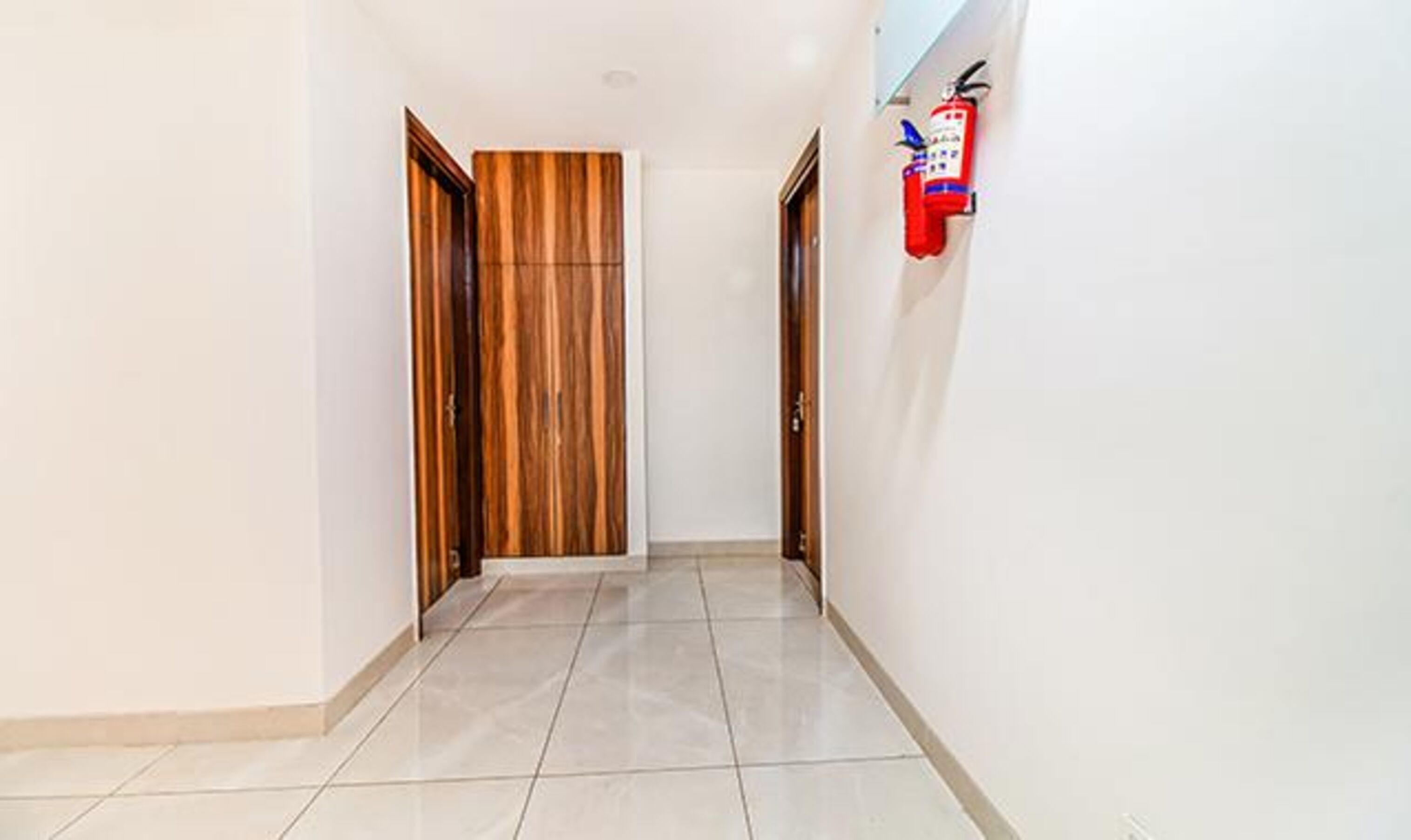 Un pasillo con una puerta de madera, un extintor en la pared y suelo de baldosas.