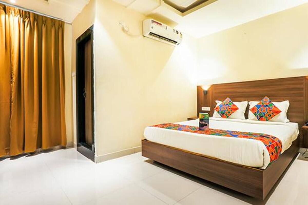 A hotel room with a wooden headboard, a bed with colorful bedding, a nightstand with a lamp, and an air conditioning unit on the wall.