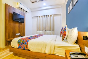 A hotel room with a wooden bed frame, a flat-screen TV mounted on the wall, a bedside table with a telephone, and a window with curtains.