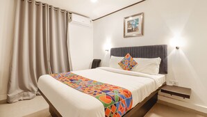 Habitación de hotel con cama, mesita de noche, lámpara, equipo de aire acondicionado, un cuadro en la pared y una ventana con cortinas.