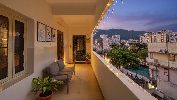 A balcony with a sofa, potted plants, and string lights. A cityscape with buildings and a river is visible in the background. - Fabhotel Aditya Yatri Nivas (Tirupati)