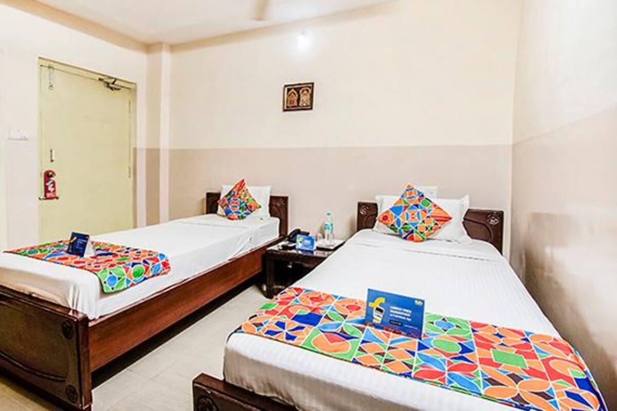 Quarto de hotel com duas camas de solteiro, cada uma com um edredom e almofadas de padrão geométrico colorido. Há uma pequena mesa de cabeceira com uma garrafa de água e um cartão sobre ela. O quarto tem um design simples e limpo, com uma porta e uma pequena moldura com uma imagem na parede.