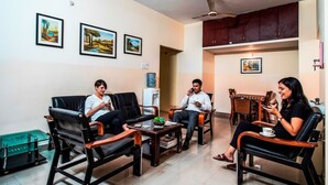 A living room with four black leather chairs, a coffee table, and a dining table with chairs. There are framed pictures on the wall and a ceiling fan. - Fabhotel Aditya Yatri Nivas (Tirupati)