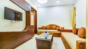 A modern living room with a flat-screen TV mounted on the wall, a wooden accent wall, a sofa with cushions, and a glass coffee table. - FabHotel Nayyar Inn (Amritsar)