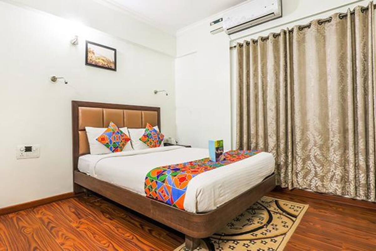 A hotel room with a wooden floor, a bed with a colorful bedspread, a headboard, an air conditioning unit, a curtain, and a small rug.