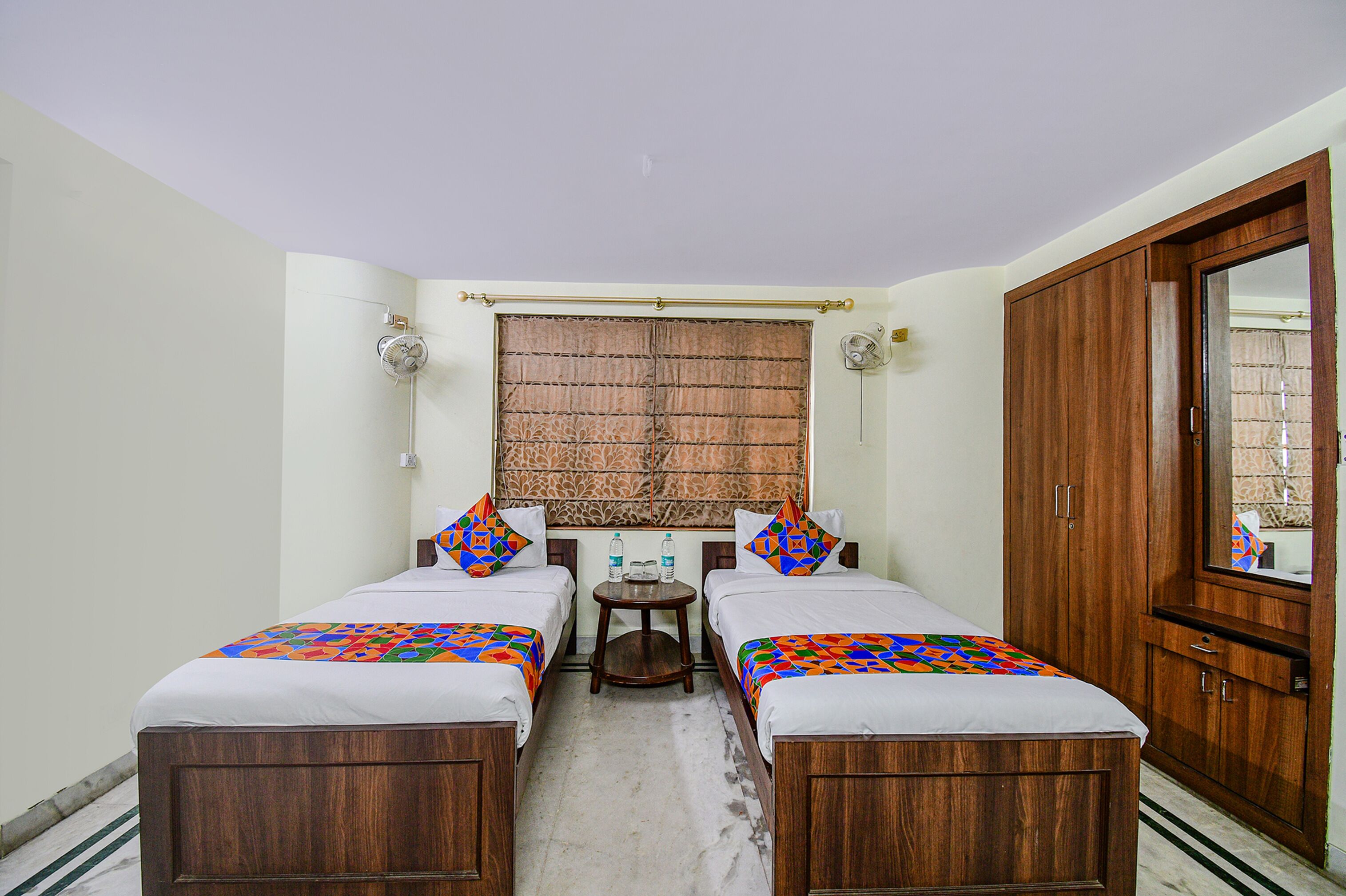 A room with two wooden beds, a small round table with water bottles, a wooden wardrobe, and a window with a patterned curtain.