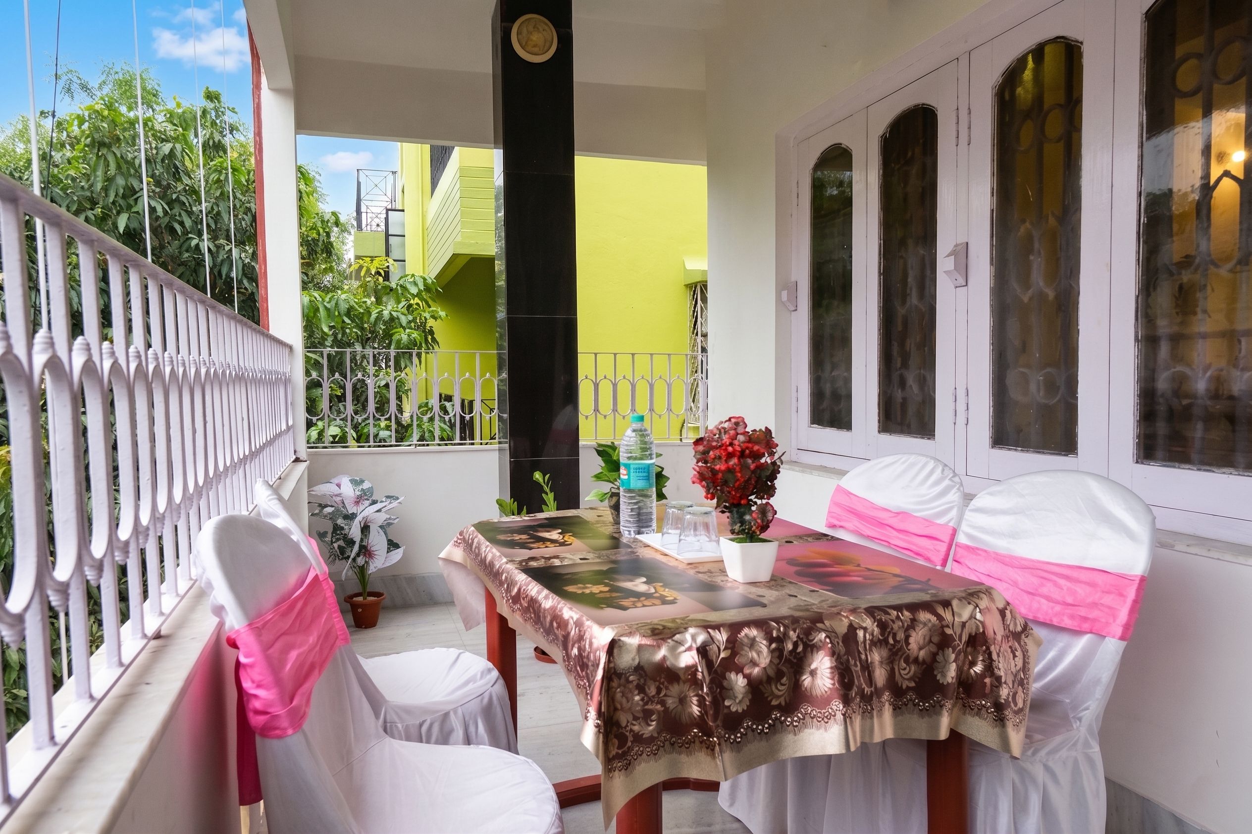Un balcón con una mesa preparada para dos, sillas adornadas con cintas rosas, una planta en maceta y una botella de agua.