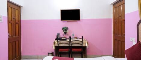 Una habitación con una cama, un escritorio con silla, un televisor montado en la pared y una pequeña planta en maceta.