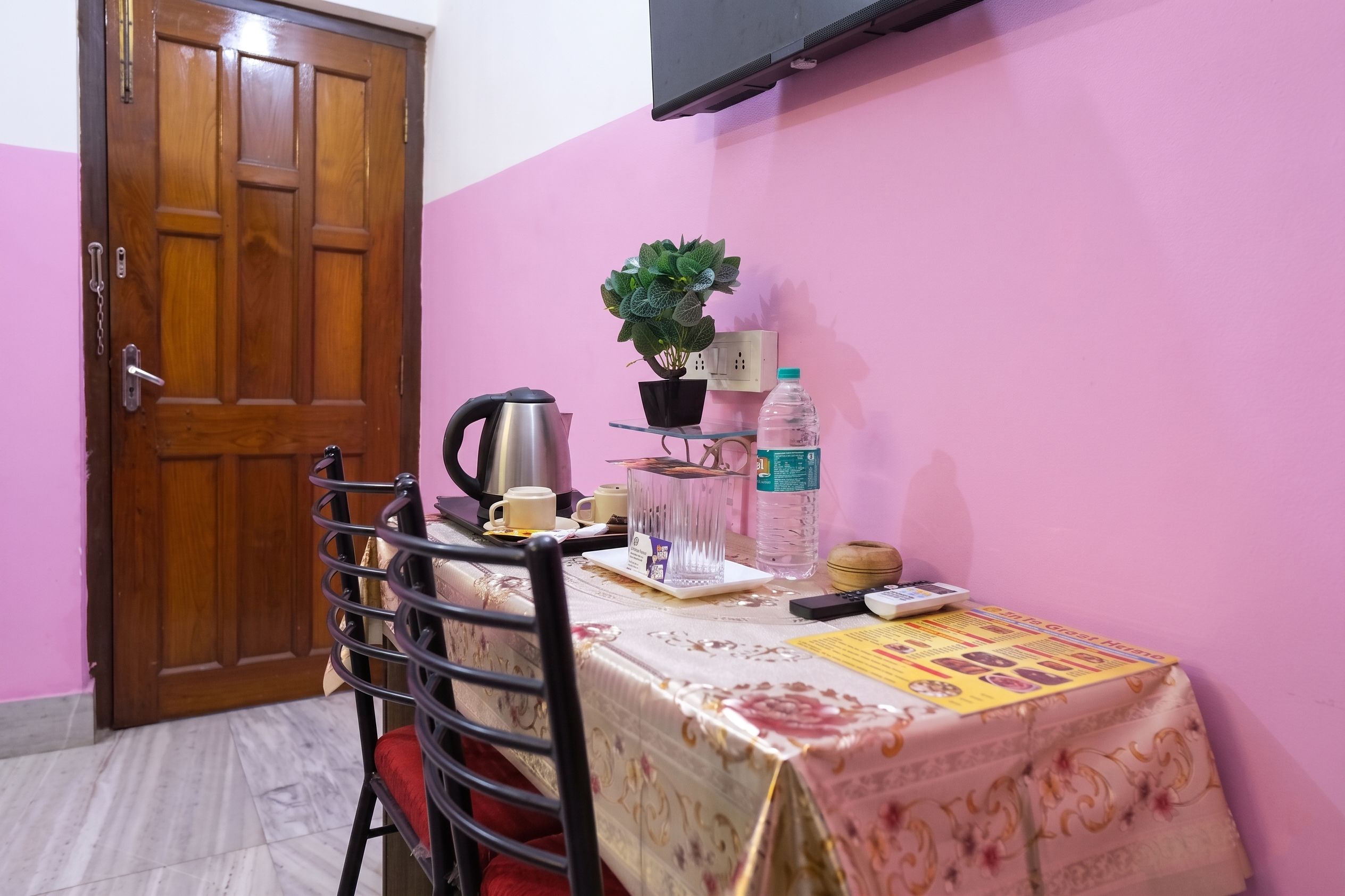 A small room with a wooden door, a table with a floral tablecloth, a chair, a potted plant, a kettle, cups, a bottle of water, a remote control, and a magazine.