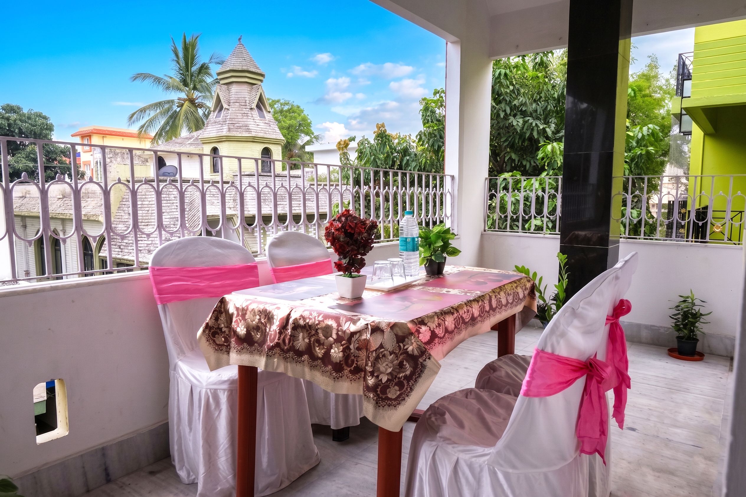 A balcony with a table set for a small gathering, surrounded by white chairs with pink sashes, a potted plant, and a bottle of water.