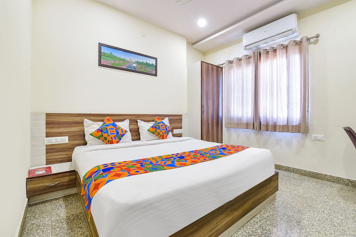 A hotel room with a bed, a nightstand, an air conditioning unit, a window with curtains, and a wall-mounted picture.