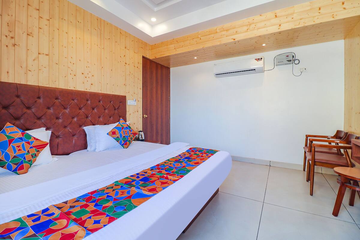 A hotel room with a large bed, wooden headboard, and a colorful bed runner. There is a wooden chair and a small table in the corner.