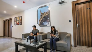 A lobby with a glass coffee table, two grey sofas, a framed picture of the Eiffel Tower, and a wooden door with a number plate. - Fabhotel F9 Sector 58 (Noida)