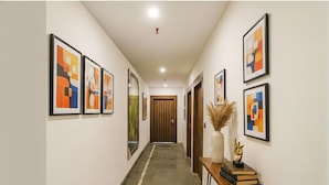 A hallway with framed abstract art on the walls, a wooden door at the end, and a small table with books and a vase of dried plants. - Fabhotel F9 Sector 58 (Noida)