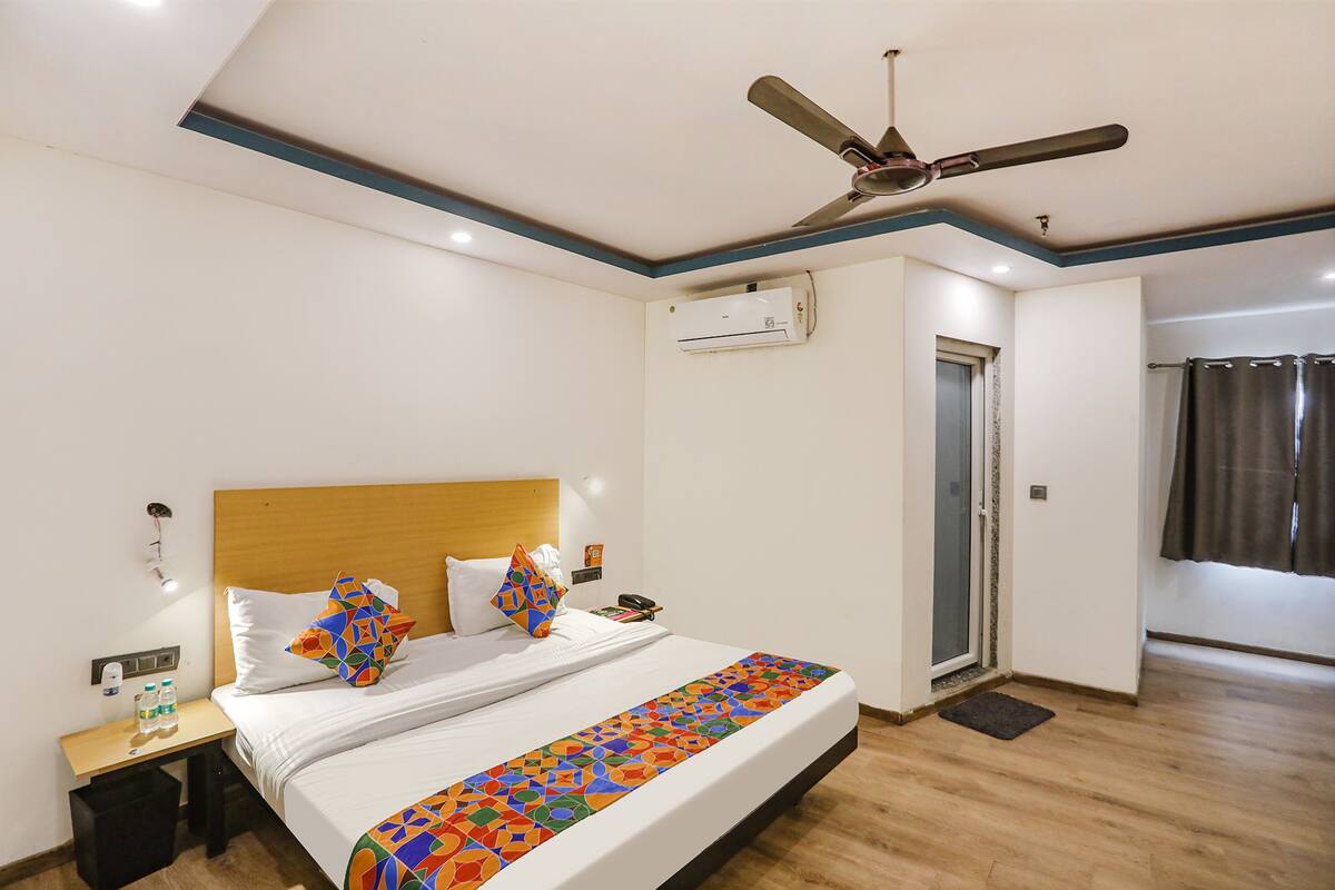 A hotel room with a bed, a nightstand, a ceiling fan, an air conditioning unit, a door, and a window with curtains.