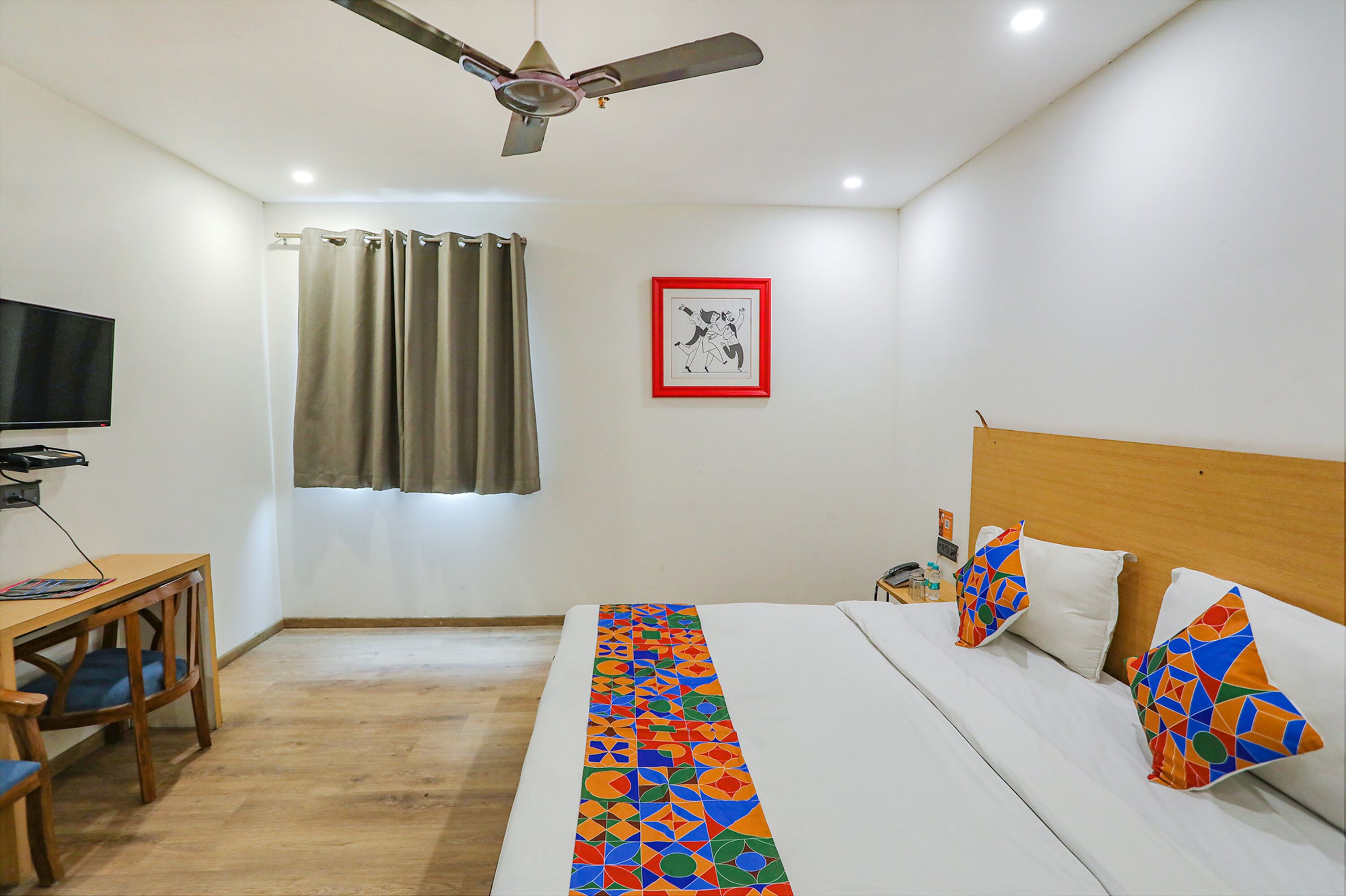 A hotel room with a bed, a desk, a chair, a television, a ceiling fan, and a window with curtains.