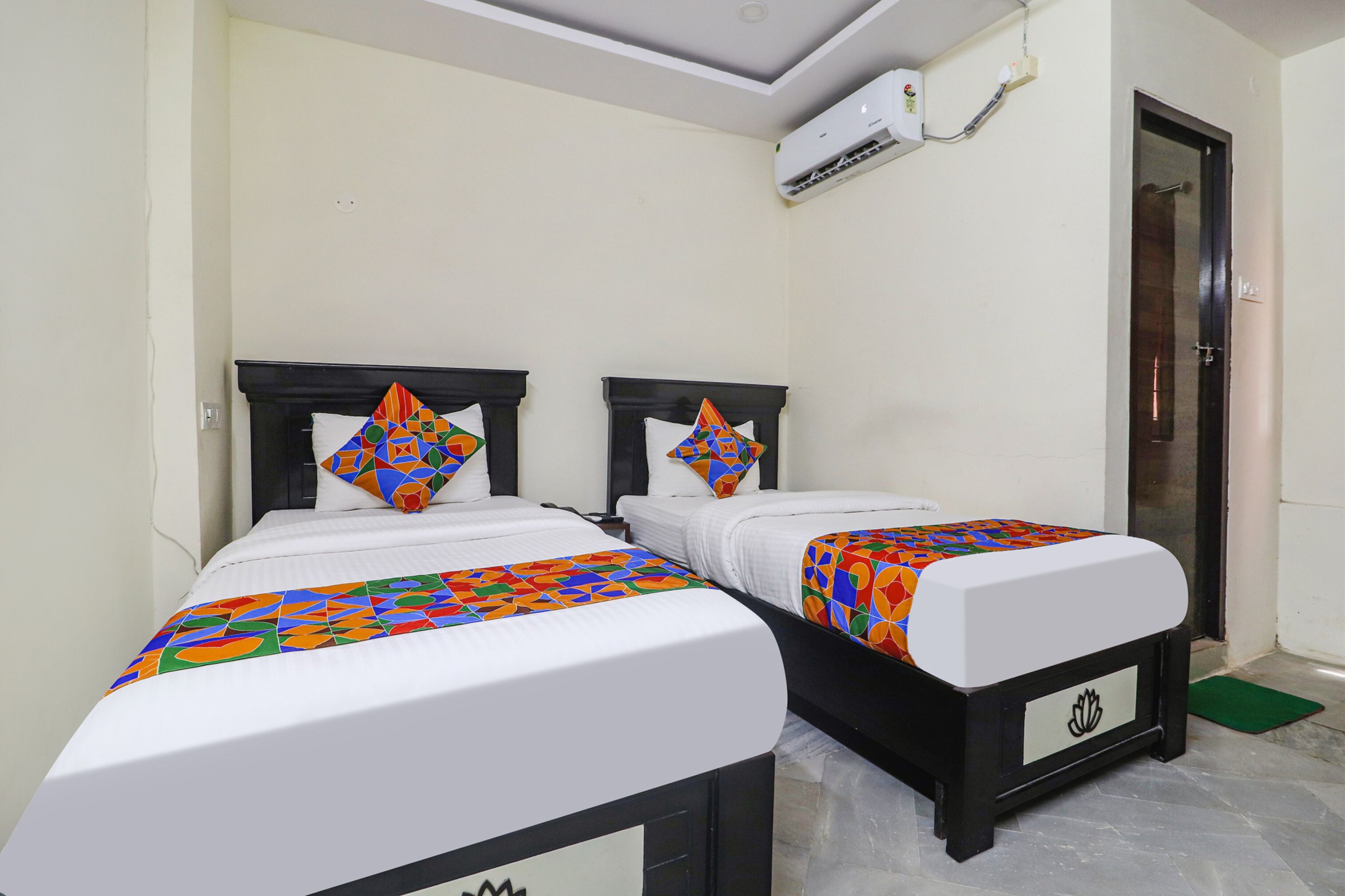 Two twin beds with matching headboards and colorful bedding, a white air conditioning unit, and a door with a green mat.