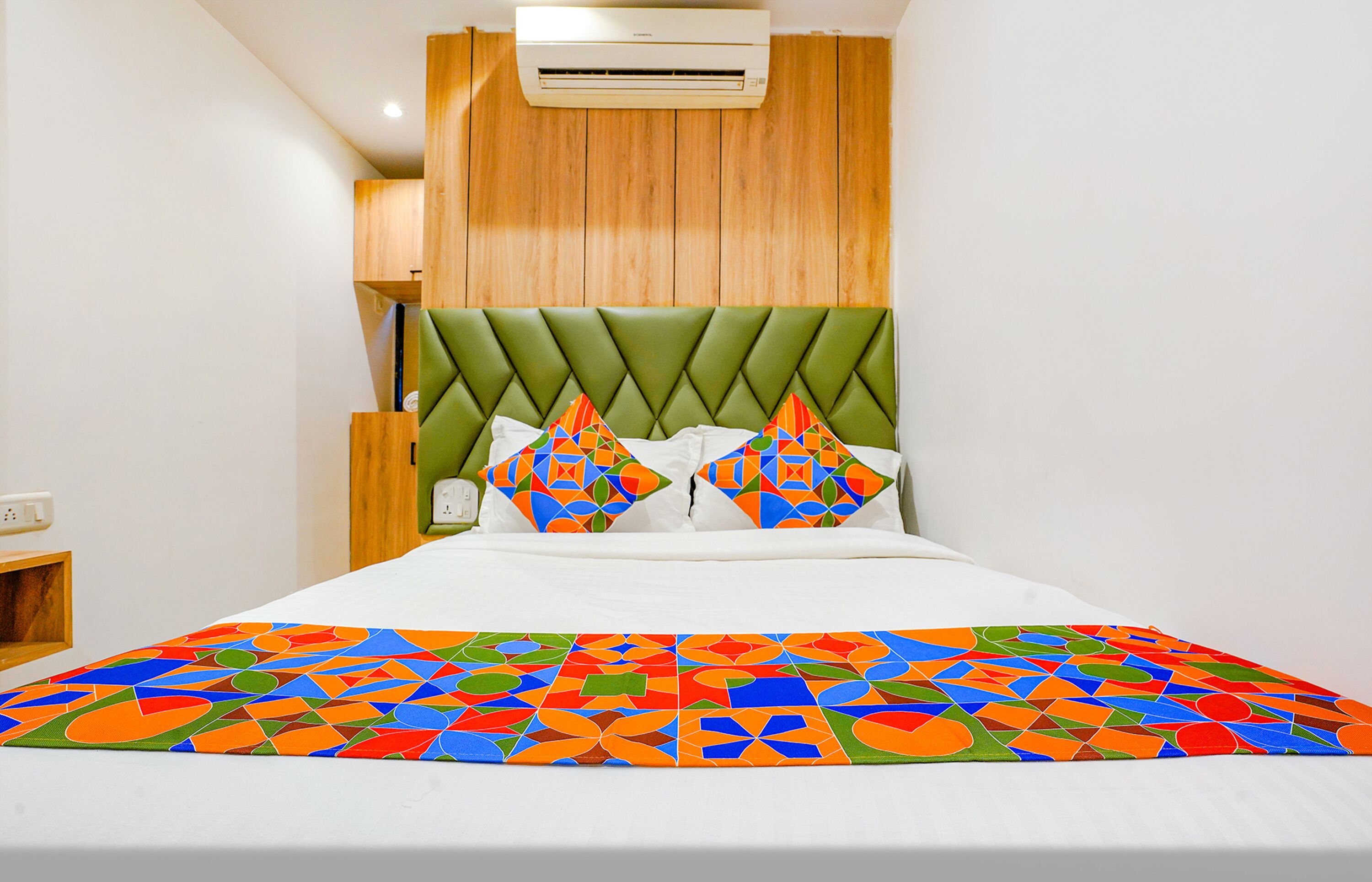 Une chambre d’hôtel moderne avec une tête de lit recouverte d’un tissu vert, une couvre-lit aux motifs géométriques colorés et un oreiller blanc orné d’un motif géométrique.