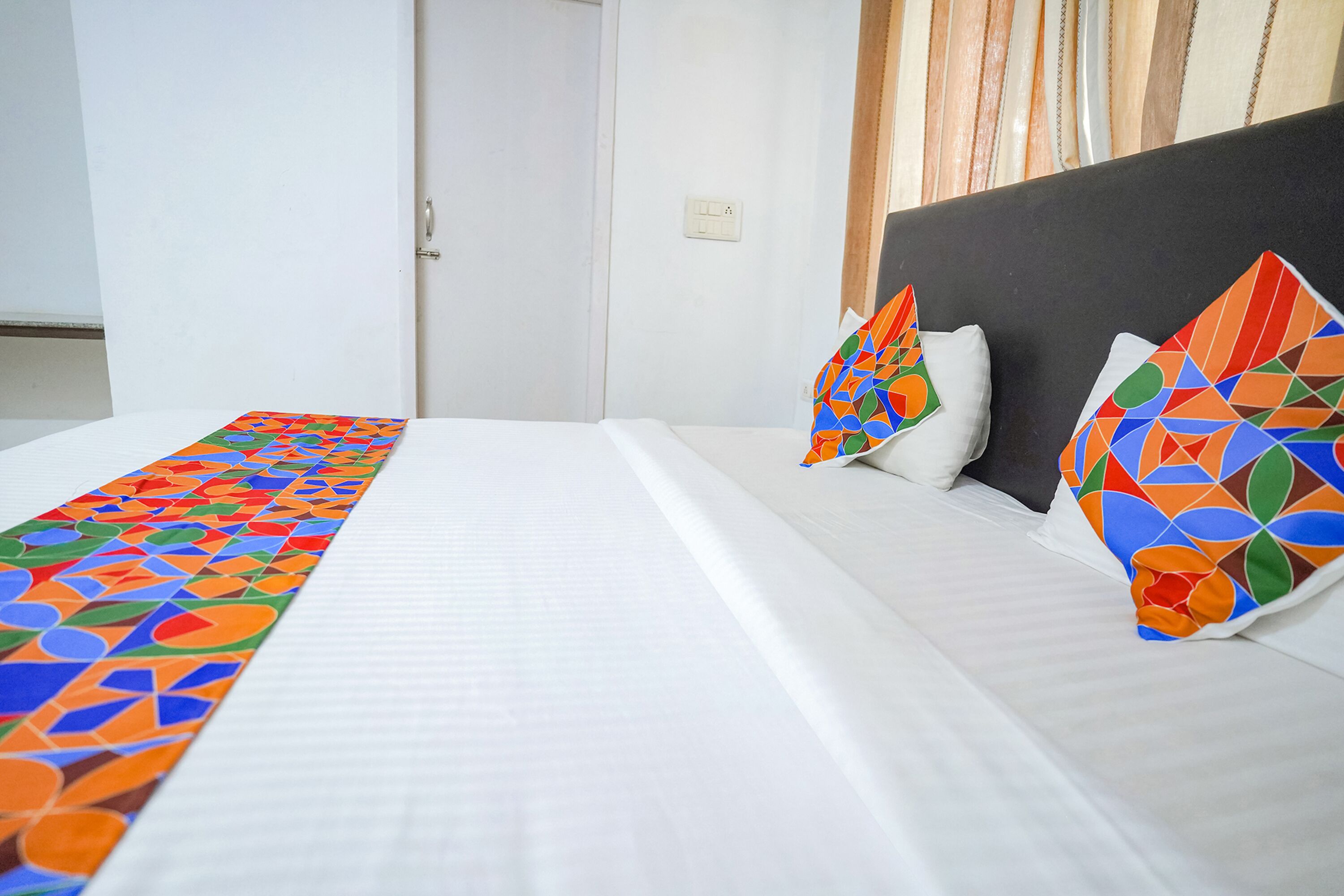 A hotel room with a neatly made bed, a colorful runner, and two decorative pillows.