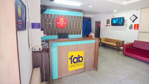 Zona de recepción de hotel con un mostrador de recepción que dice "fab hotels", un cartel de "KB Residency", un televisor y un sofá rojo.