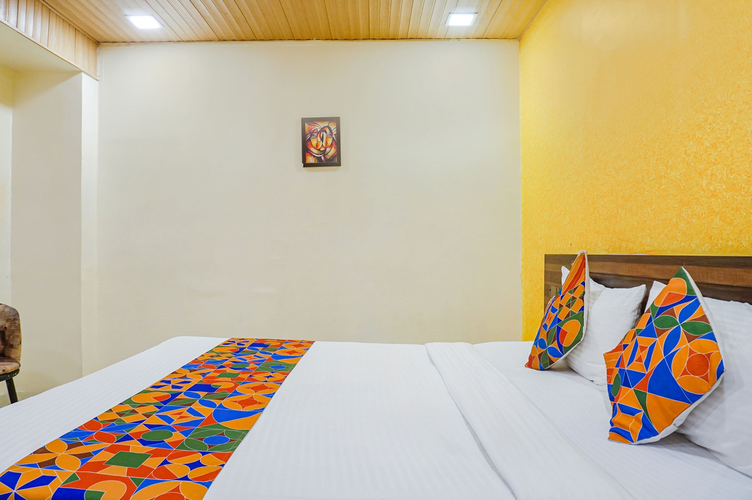 A hotel room with a bed, a colorful bed runner, a painting on the wall, and a chair.