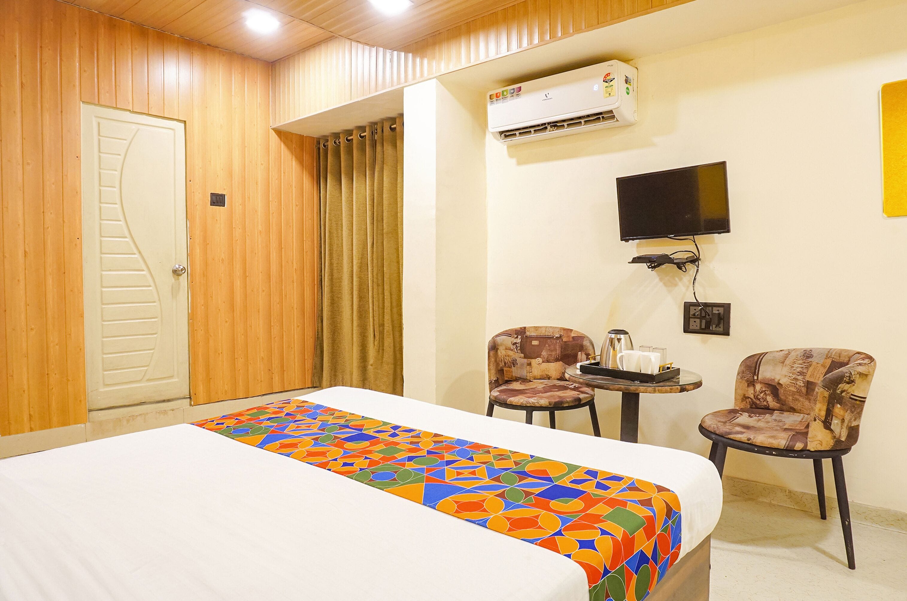 A hotel room with a bed, a TV, two chairs, and an air conditioning unit.