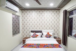 A neatly arranged bedroom with a patterned wallpaper, a bed with colorful geometric pillows, a bedside table with a phone, and an air conditioning unit on the wall.