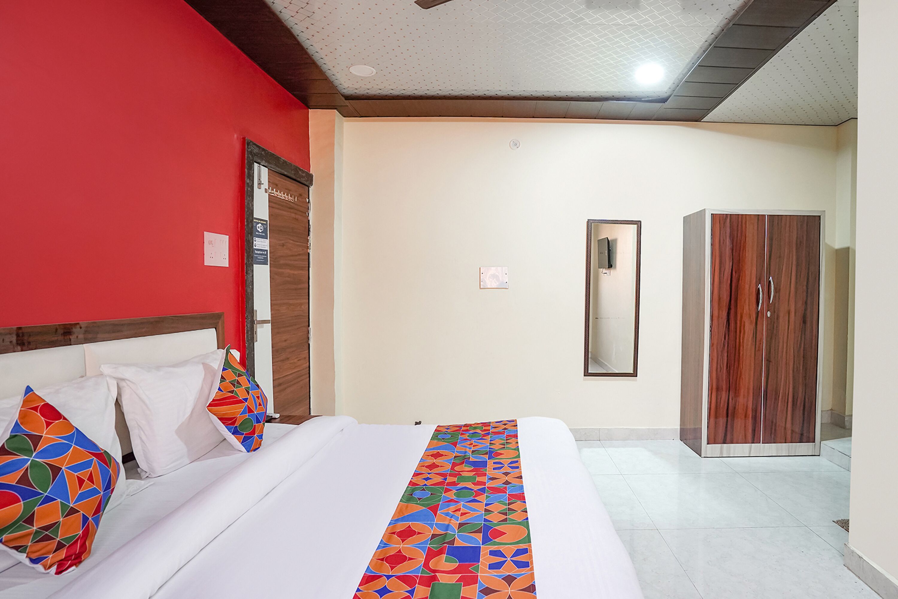 A hotel room with a bed, a red wall, a wooden wardrobe, and a mirror.