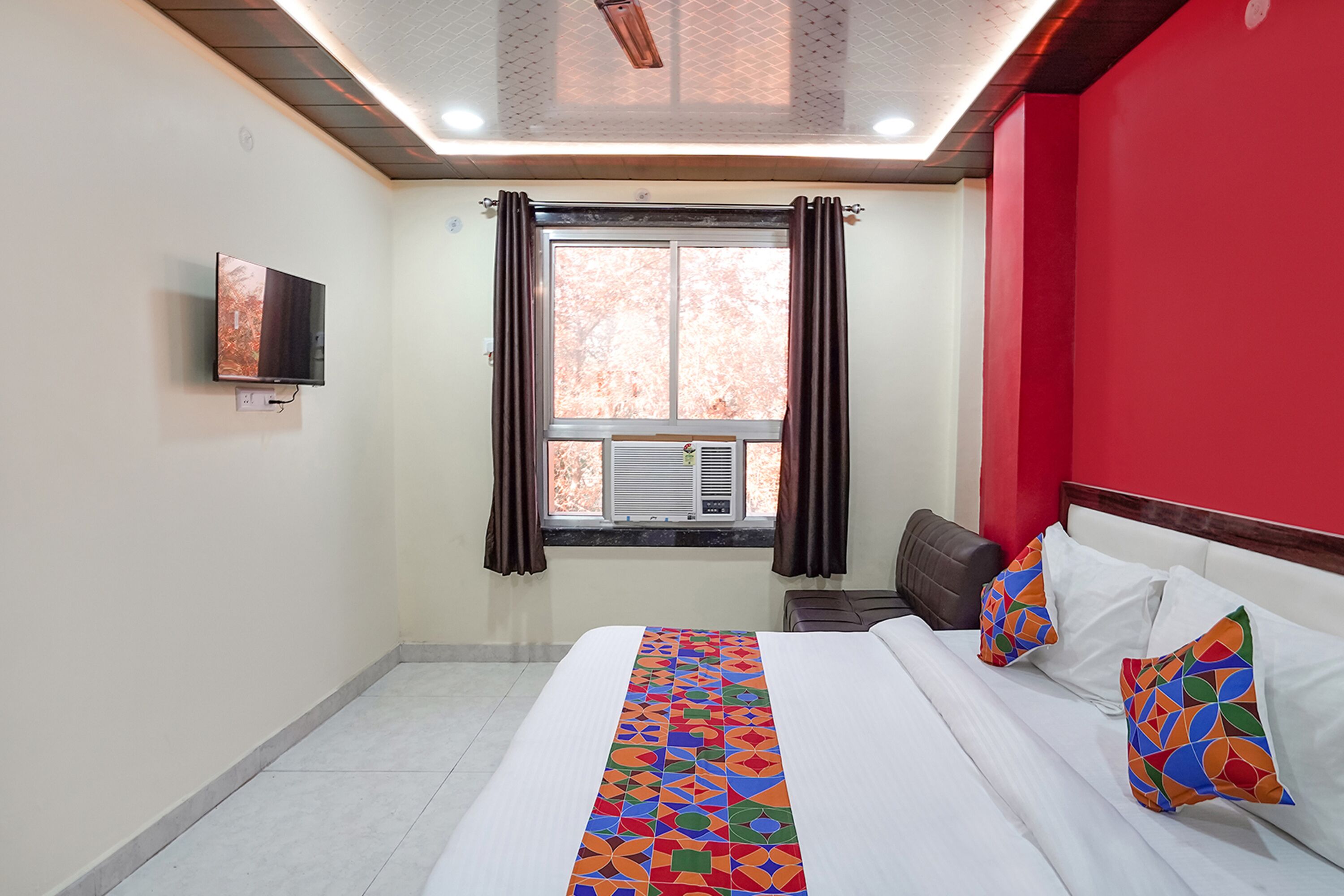 Una habitación de hotel con cama, televisor montado en la pared, ventana con cortinas y una pared de acento roja.