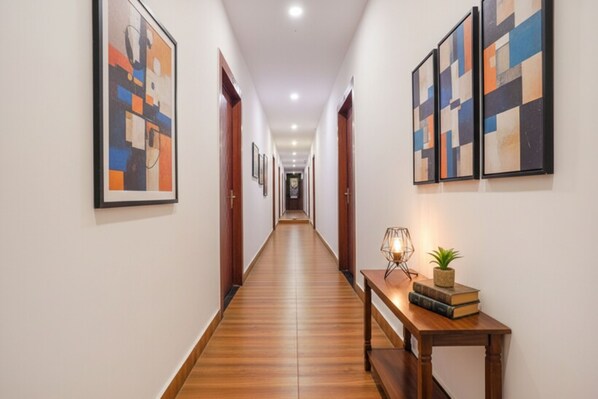 A hallway with wooden flooring, framed abstract art on the walls, and a wooden bench with books and a lamp. - Fabexpress Gauri Shree (Bhopal)