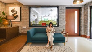 Een vrouw zit op een smaragdgroene bank in een moderne kamer met een stenen muur, een groot raam en een houten deur.