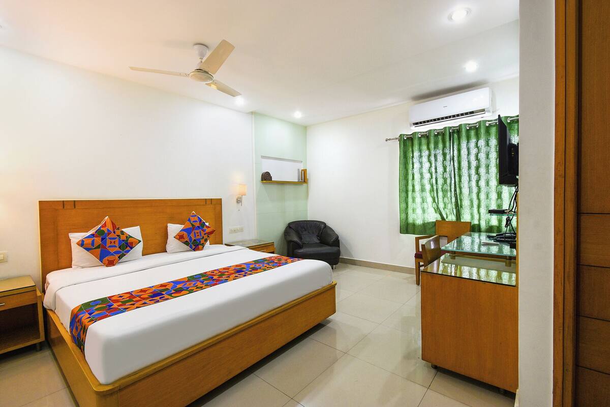 A hotel room with a bed, a desk, a chair, a ceiling fan, an air conditioning unit, and a green curtain.