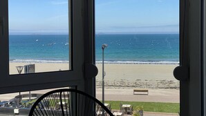 Vista desde una ventana hacia una playa, cielo despejado, un banco y un farol.