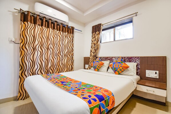 A neatly made bed with a colorful geometric-patterned blanket, a wooden nightstand with a drawer, a wall-mounted air conditioner, and a window with a view of trees.