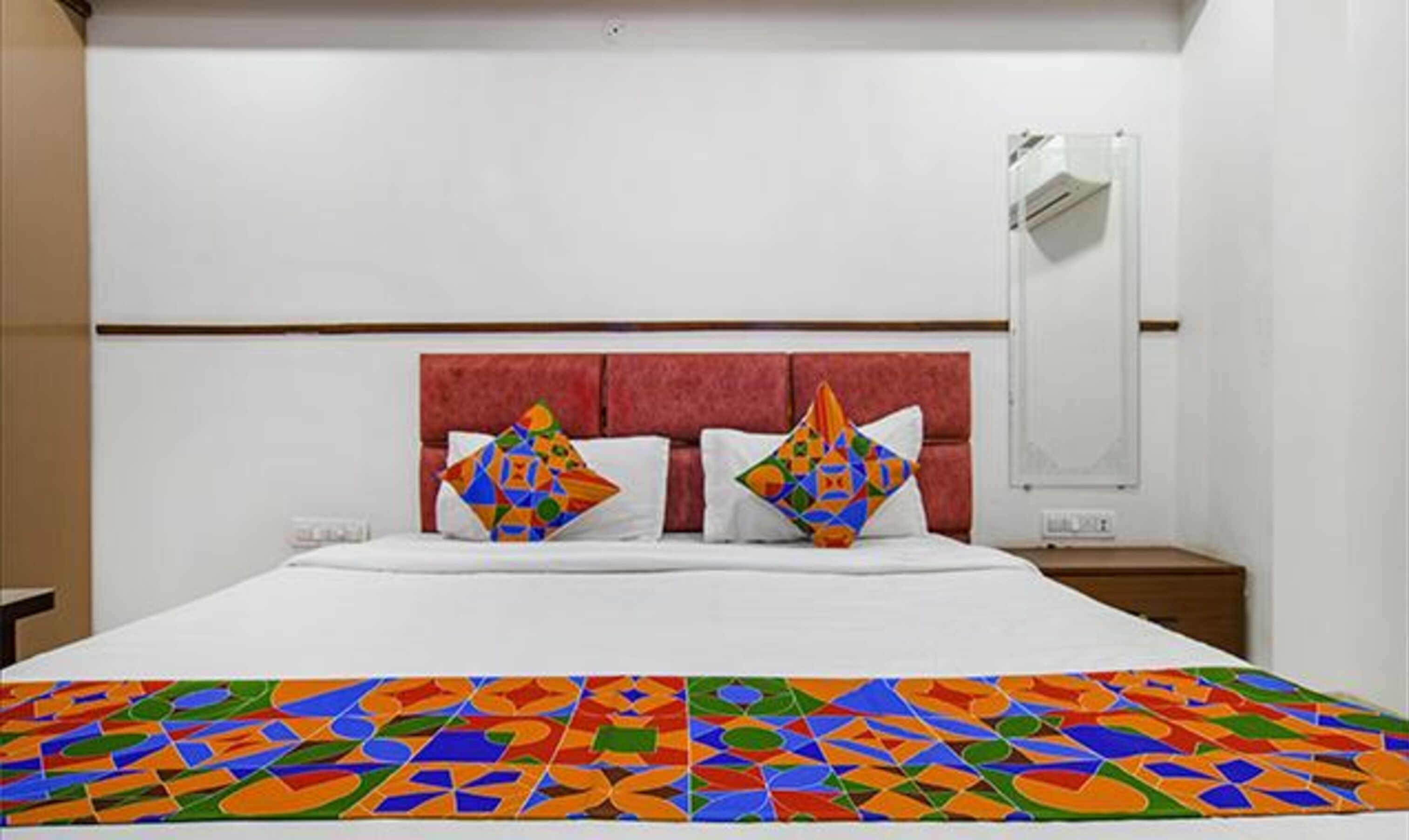 A hotel room with a bed, two colorful pillows, a nightstand, and a mirror.