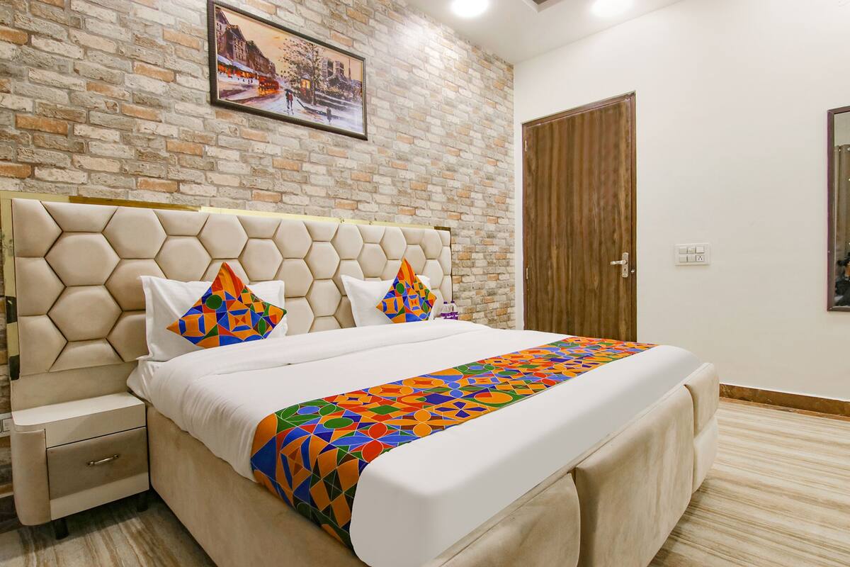 Una habitación de hotel con una pared de ladrillo, una puerta de madera, una cama con colcha de estampado geométrico y una mesita de noche con cajón.