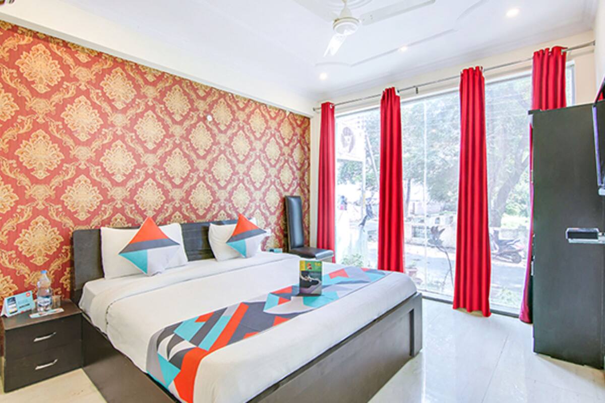 Un dormitorio con papel tapiz estampado, una cama con ropa de cama colorida, una mesita de noche, un televisor y un ventanal corredero con cortinas rojas.